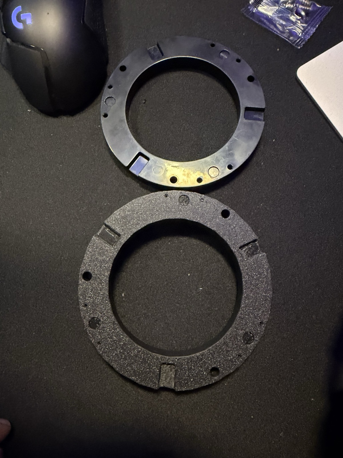 Two puck mounting rings on a desk: the stock injection-molded ring above, a black printed replacement below.